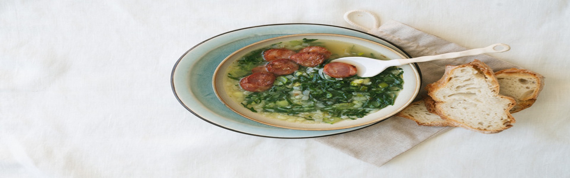 Caldo Verde Soup with greens and chopped chorizo on the top in ceramic bowl over ceramic plate on cotton bag with ceramic spoon served with pieces of bread. Top View